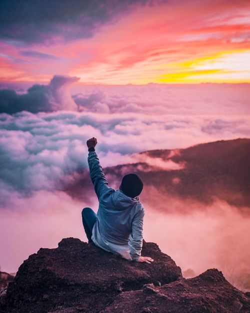 man-sitting-on-mountain-cliff-facing-white-clouds-rising-one-hand-at-golden-hour-bH7kZ0yazB0.jpg